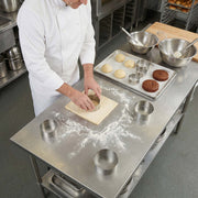 Baker in a professional kitchen rolling out dough on a stainless steel counter.