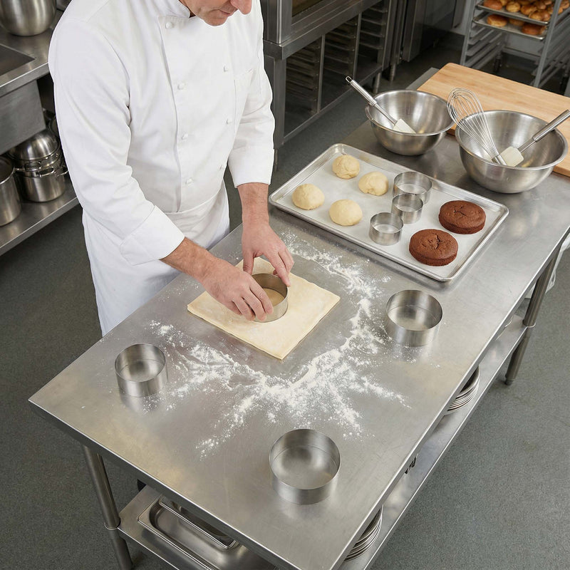 Baker in a professional kitchen rolling out dough on a stainless steel counter.