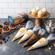 A high-angle, studio-lit shot of a kitchen countertop featuring baking supplies. In the foreground, four clear disposable pastry bags filled with frosting—three with white cream and one with chocolate ganache—are arranged diagonally on a dark grey marble surface. Each bag is branded with the "EXNOX" logo. To the left, a wire cooling rack holds several small fruit tarts topped with fresh raspberries and blackberries. In the background, a wooden cake stand displays four frosted cupcakes, positioned next to a 