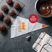 A top-down, flat-lay studio shot on a dark grey slate surface. On the left, a metal wire cooling rack holds several freshly baked chocolate cupcakes in blue liners. In the center, a large, empty, transparent 20” long plastic piping bag with yellow branding text lies diagonally. To the right, a stainless steel mixing bowl is filled with rich chocolate frosting. Nearby, a small metal piping tip sits on the surface, and a white silicone spatula rests on a folded white kitchen towel with black stripes. The ligh