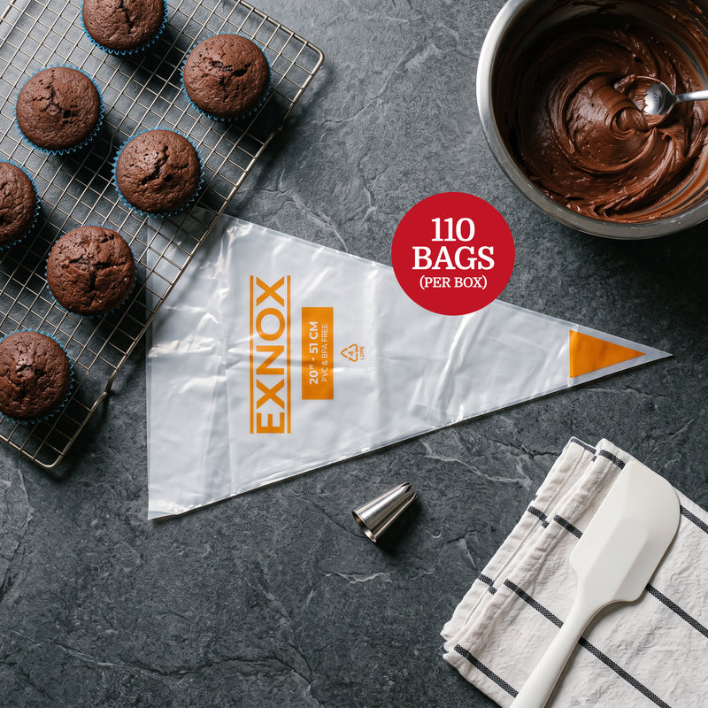 A top-down, flat-lay studio shot on a dark grey slate surface. On the left, a metal wire cooling rack holds several freshly baked chocolate cupcakes in blue liners. In the center, a large, empty, transparent 20” long plastic piping bag with yellow branding text lies diagonally. To the right, a stainless steel mixing bowl is filled with rich chocolate frosting. Nearby, a small metal piping tip sits on the surface, and a white silicone spatula rests on a folded white kitchen towel with black stripes. The ligh